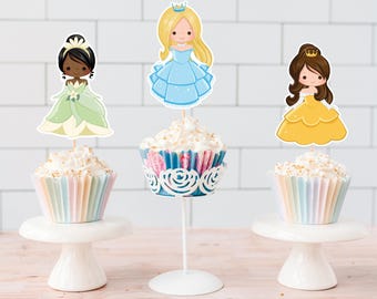 May include: Three cupcakes decorated with princess themed toppers. The cupcakes are on white cake stands. The princess toppers are of Tiana, Cinderella, and Belle.