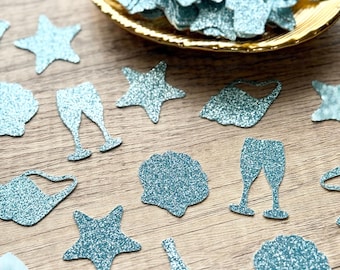 May include: Light blue glitter confetti in the shapes of champagne flutes, bottles, stars, and seashells. A collection of these sparkling paper cutouts is contained within a small, oval-shaped gold dish, while others are scattered around.