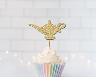 May include: A gold glitter magic lamp cupcake topper attached to a thin wooden pick, inserted into a white frosted cupcake with a pastel-colored pleated paper liner. The lamp shape features a curved spout and a handle, creating a decorative accent.