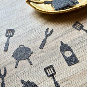 May include: A collection of black glittery paper cutouts shaped like barbecue tools, including spatulas, forks, condiment bottles, and round grills. Some pieces are scattered across the surface, while others are inside a gold leaf-shaped dish.