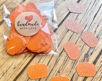 May include: A clear plastic bag filled with orange glitter pumpkin-shaped paper cutouts, featuring a round brown sticker with a red heart and the text handmade WITH LOVE. Several loose orange glitter pumpkin cutouts are scattered nearby.