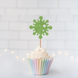 May include: A cupcake with pastel-colored paper and white frosting, topped with a glittery green decoration. The decoration is shaped like a stylized virus or snowflake, attached to a wooden pick.