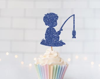 May include: A dark blue glitter cupcake topper shaped like a silhouette of a person sitting and holding a fishing rod with a fish on the line. The topper is inserted into a cupcake with a pastel-colored pleated paper liner.