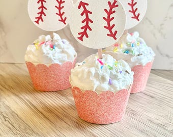May include: Three cupcakes with white frosting and colorful sprinkles, each featuring a round white paper topper with a red baseball stitch design. The cupcakes are held in glittery coral-pink scalloped wrappers, creating a festive display.