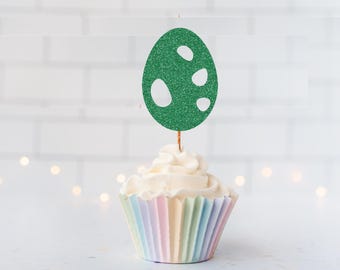 May include: A green glittery egg-shaped cupcake topper with irregular white cut-out spots, attached to a thin wooden pick, inserted into a cupcake with white frosting in a pastel-striped paper liner. The topper features a textured finish.