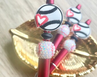 May include: A set of three pens with matte red barrels, featuring a baseball-themed topper with a white baseball and a red heart. The pens are decorated with a white, iridescent, textured bead resembling a baseball and smaller red spacers.