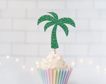 May include: A green glitter palm tree cupcake topper inserted into a white frosted cupcake with a pastel striped paper liner. The palm tree cutout features a textured, sparkling finish and is attached to a thin, light-colored wooden pick.