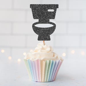 May include: A cupcake with white icing and a black glitter toilet-shaped cake topper. The cupcake case has pastel vertical stripes of pink, blue, yellow, and green. The background is a white brick wall with soft, out-of-focus lights.