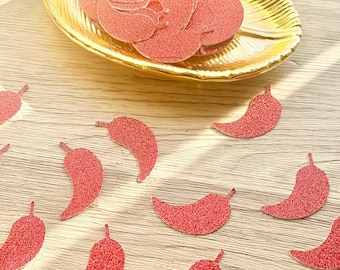 May include: A collection of small, red, glittery chili pepper-shaped cutouts scattered around a gold, leaf-shaped decorative dish. The chili pepper shapes are textured with a sparkling finish and are arranged both inside the dish and on the surface.