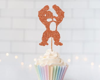 May include: A copper-colored glitter cupcake topper shaped like a muscular gorilla silhouette with arms raised, attached to a thin wooden pick, inserted into a cupcake with white frosting and a pastel-striped paper liner.