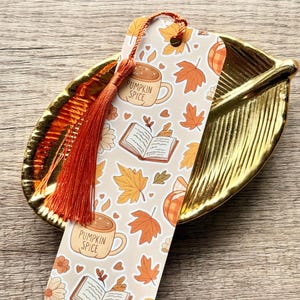 May include: A rectangular paper bookmark with a light beige background featuring a repeating pattern of orange maple leaves, open books, and mugs labeled PUMPKIN SPICE. The bookmark has a vibrant orange silk tassel attached to the top center.