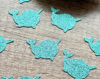May include: A collection of small, flat, glittery teal narwhal-shaped cutouts. Each cutout features a rounded body, a small tail fluke, and a single pointed tusk extending from the head. The surface of each narwhal shape is covered in a fine, sparkling teal glitter texture.