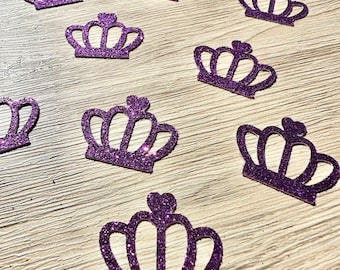May include: A collection of ten purple glitter crown-shaped cutouts. Each cutout features a five-point design with a small heart at the top center. The crowns are scattered across the frame, displaying a textured, sparkling surface finish.