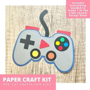 May include: A gray paper craft kit shaped like a video game controller with a blue outline. The controller has red, yellow, green, and orange buttons, a black plus sign, and a red triangle. Text on the image reads "PAPER CRAFT KIT" and "PRE-CUT CRAFTS FOR KIDS."