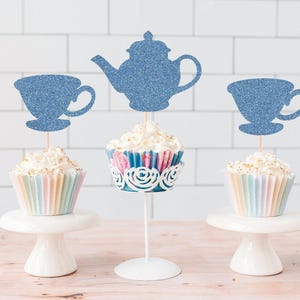 May include: Cupcakes with blue glitter teapot and teacup toppers. The cupcakes have pastel-coloured wrappers and are displayed on white cake stands. The background is a white tile wall.