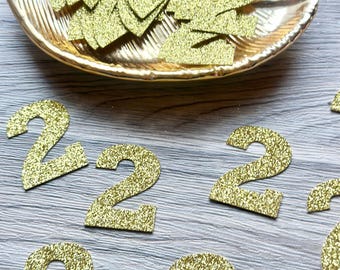 May include: A collection of gold glitter number two cutouts scattered on a surface, with several additional gold glitter number two cutouts resting inside a gold-colored, leaf-shaped decorative dish with a textured, ribbed surface.
