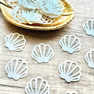 May include: A collection of light blue seashell-shaped confetti scattered on a wooden surface, with some arranged in a gold-colored leaf-shaped dish. The confetti has a glittery texture, adding a touch of sparkle.