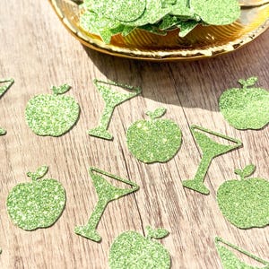 Peut inclure: Une collection de confettis vert vif à paillettes en forme de pommes et de verres à martini. Certaines pièces sont éparpillées sur la surface, tandis que d'autres sont contenues dans un plat décoratif doré en forme de feuille.