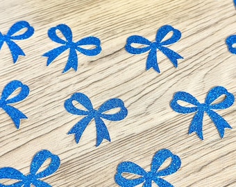 May include: A collection of small, blue glittery bow-shaped cutouts scattered across a surface, with a gold-colored dish containing more of the same blue bow cutouts positioned at the top of the frame.
