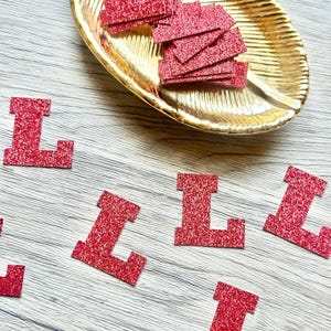 May include: A collection of red glitter letter L cutouts scattered across the surface, with a stack of identical red glitter letter L shapes resting inside a gold-colored, leaf-shaped decorative tray with a ribbed texture.