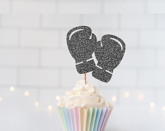 May include: A black glittery boxing glove cupcake topper attached to a thin wooden pick, inserted into a cupcake with white frosting and a pastel-colored pleated paper liner. The topper features two stylized boxing gloves side by side.