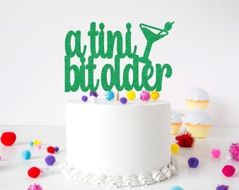 May include: A green glitter cake topper featuring the text "a tini bit older" in a cursive font, accompanied by a martini glass icon. The topper is positioned on top of a plain white frosted cake decorated with small, colorful pom-poms.