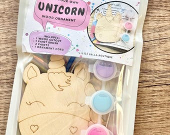May include: A paint your own unicorn wood ornament kit in a clear plastic pouch. The kit includes one wooden unicorn cutout, one paint brush, three small paint pots in purple, blue, and pink, and one ornament cord. The label reads Paint Your Own Unicorn Wood Ornament and Little Bella Boutique.
