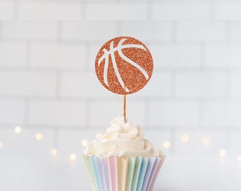 May include: A cupcake topper featuring a circular orange glitter basketball design with white lines, attached to a thin wooden pick, inserted into a white frosted cupcake with a pastel-colored pleated paper liner.