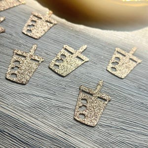 May include: Multiple small, gold-colored glittery cutouts shaped like bubble tea cups with straws. Each cutout features a distinct cup silhouette with a straw extending from the top, covered in a textured, sparkling gold finish that reflects light.