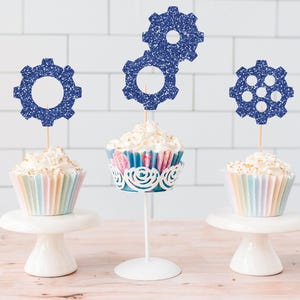 May include: Three cupcakes with white frosting and blue glitter gear toppers. The cupcakes sit on white cake stands and a decorative metal stand. The cupcake liners are pastel colors.