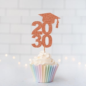 May include: A copper-colored glitter cupcake topper featuring the numbers 2030 and a graduation cap icon. The topper is inserted into a white frosted cupcake with a pastel-colored pleated paper liner, creating a festive graduation celebration display.