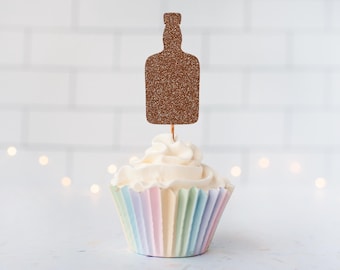 May include: A cupcake topper shaped like a liquor bottle with a brown glitter finish, attached to a thin wooden pick. The topper is inserted into a cupcake with white frosting and a pleated paper liner featuring pastel stripes of pink, yellow, and blue.