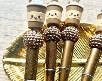 May include: A set of four pens with brown glitter barrels and silver accents. Each pen features a topper shaped like a smiling coffee cup with a lid and straw. Below the cup is a decorative bead cluster with a metallic brown finish.
