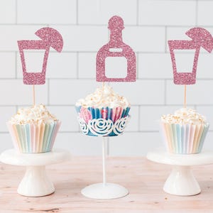 May include: Three cupcakes topped with white frosting and pink glitter cardstock toppers. Two toppers are shaped like cocktail glasses with lime wedges, and the center topper is shaped like a tequila bottle. Each is mounted on a thin wooden stick.