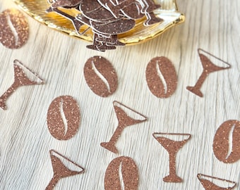 May include: A collection of copper-colored glitter confetti shaped like coffee beans and martini glasses. Some pieces are contained within a gold-toned leaf-shaped dish, while others are scattered individually across the surface.