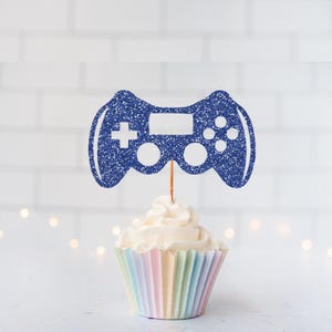 May include: A cupcake with a blue glittery video game controller topper. The cupcake has a pastel-colored liner and is topped with white frosting. The background is a white brick wall with soft, out-of-focus lights.