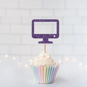 May include: A cupcake topped with a purple glitter computer monitor cake decoration. The cupcake has a swirl of white icing and a pastel-striped paper case. The background is a white brick wall with soft, blurred lights.