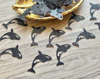 May include: A collection of black glittery orca whale cutouts scattered across a surface, with several pieces resting inside a gold-colored, leaf-shaped dish. Each cutout features the distinct silhouette of an orca in a jumping pose.