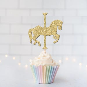 May include: A cupcake topped with a gold glitter carousel horse cake topper. The cupcake has a pastel-colored liner and white frosting. The background is a white brick wall with soft, out-of-focus lights.