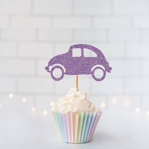 May include: A purple glitter car-shaped cupcake topper attached to a thin wooden pick, positioned vertically in the center of a white frosted cupcake with a pastel-colored pleated paper liner. The car silhouette features two circular wheels.