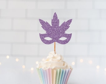 May include: A purple glittery masquerade mask cupcake topper shaped like a leaf, attached to a thin wooden pick, inserted into a white frosted cupcake with a pastel-colored pleated paper liner. The topper features two eye cutouts.
