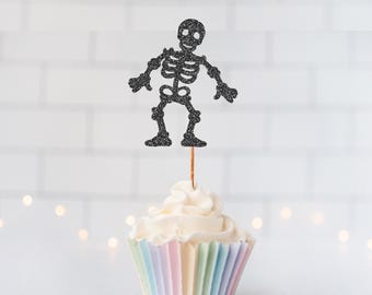 May include: A black glitter skeleton cupcake topper attached to a thin wooden pick, inserted into a cupcake with white frosting and a pastel-colored pleated paper liner. The skeleton figure has a detailed rib cage, skull, and jointed limbs.