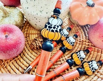 May include: A set of pens featuring orange barrels and decorative toppers shaped like stacked pumpkins with black and white cow print patterns. The pens are arranged on a woven surface alongside various decorative gourds and fruits.