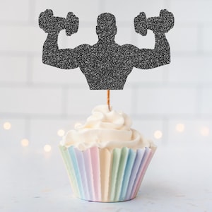 Peut inclure: Un cupcake avec un glaçage blanc et une enveloppe aux couleurs pastel. Une silhouette noire pailletée d'un haltérophile tenant des haltères est fixée au sommet du cupcake avec un bâton en bois.