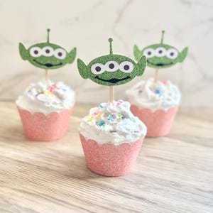 May include: Three pink cupcakes with white frosting and colorful sprinkles topped with green alien cupcake toppers. The alien toppers have three eyes and a friendly smile.