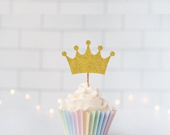 May include: A gold glitter crown cupcake topper sits atop a white frosted cupcake in a pastel striped paper liner. The crown features five points and is attached to a thin wooden pick inserted into the center of the white frosting swirl.