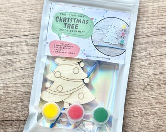May include: A paint your own Christmas tree wood ornament craft kit in a white pouch. The kit includes a wooden tree cutout, a blue paint brush, a white ornament cord, and three small paint pots in yellow, red, and green. The label reads Paint Your Own Christmas Tree Wood Ornament, Includes 1 Wood Cutout, 1 Paint Brush, 3 Paints, 1 Ornament Cord, Little Bella Boutique.