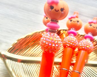 May include: Three orange pens featuring decorative toppers with a smiling pumpkin character wearing a small pink bow. Each pen includes a bright orange barrel, a sparkling orange bead, and a pink spacer bead accented with silver-toned rings.