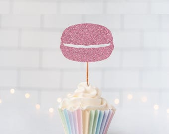 May include: A pink glittery macaron-shaped cupcake topper attached to a thin wooden stick, inserted into a cupcake with white frosting and a pastel-colored pleated paper liner. The macaron cutout features a textured, sparkling finish.