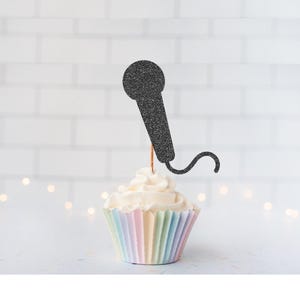 May include: A black glittery microphone-shaped cupcake topper attached to a thin wooden pick, inserted into a cupcake with white frosting and a pastel-colored pleated paper liner. The microphone design features a rounded head and a curved cord.